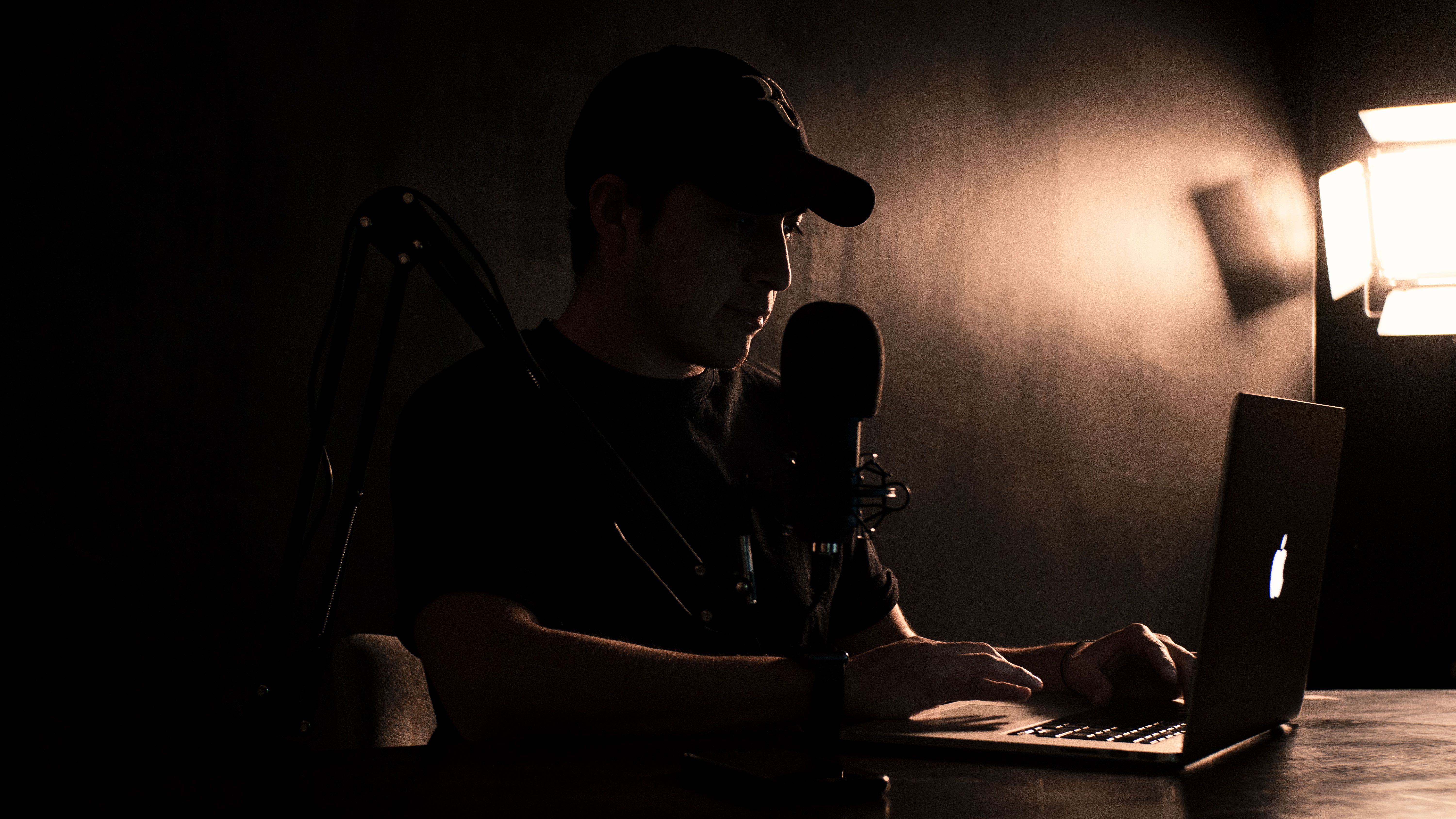 A man wearing a cap works on a laptop with a microphone setup in a dimly lit room.