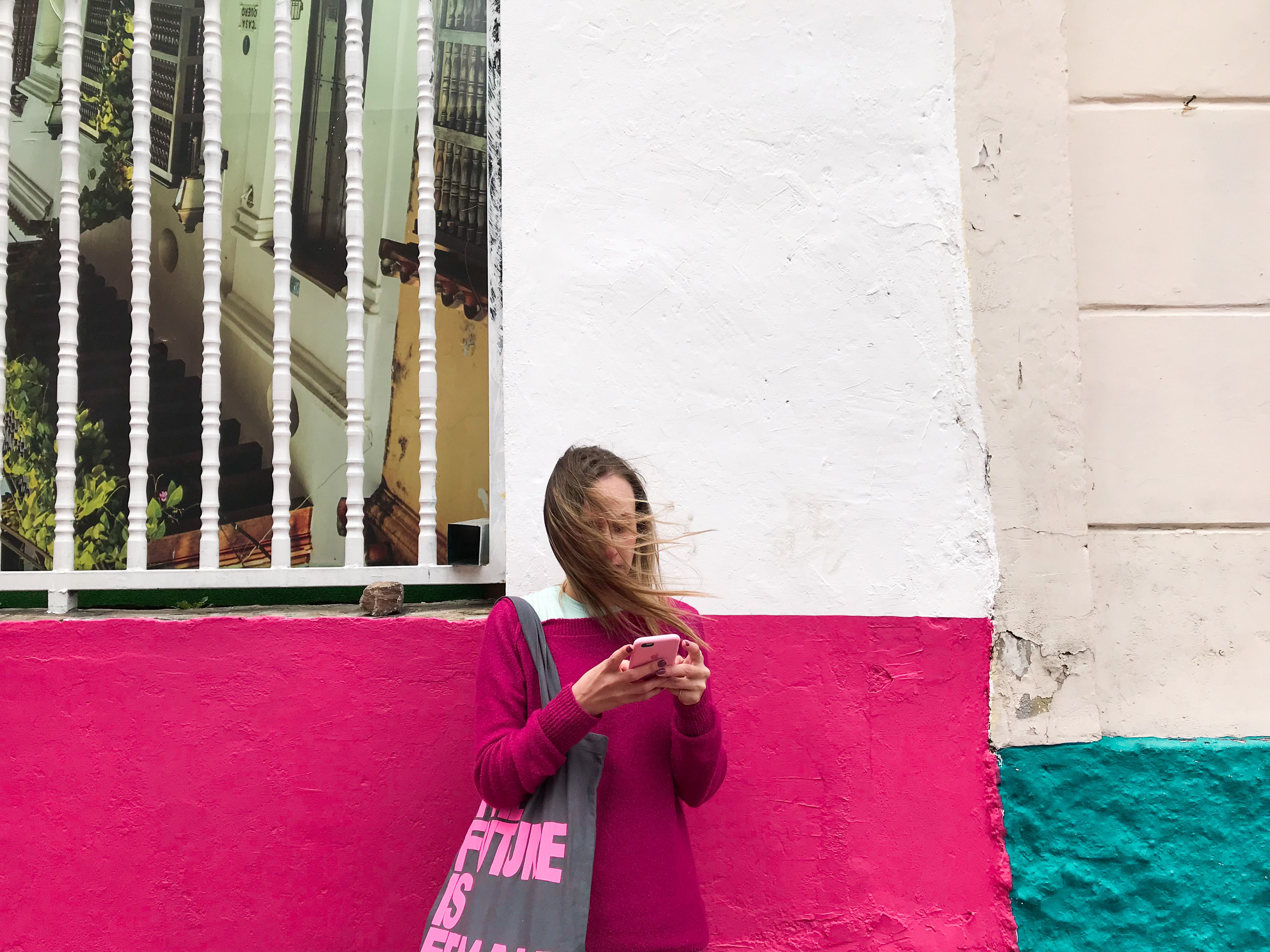 Woman with blond hair using smartphone against vibrant wall in Bogotá. Urban fashion and modern lifestyle.