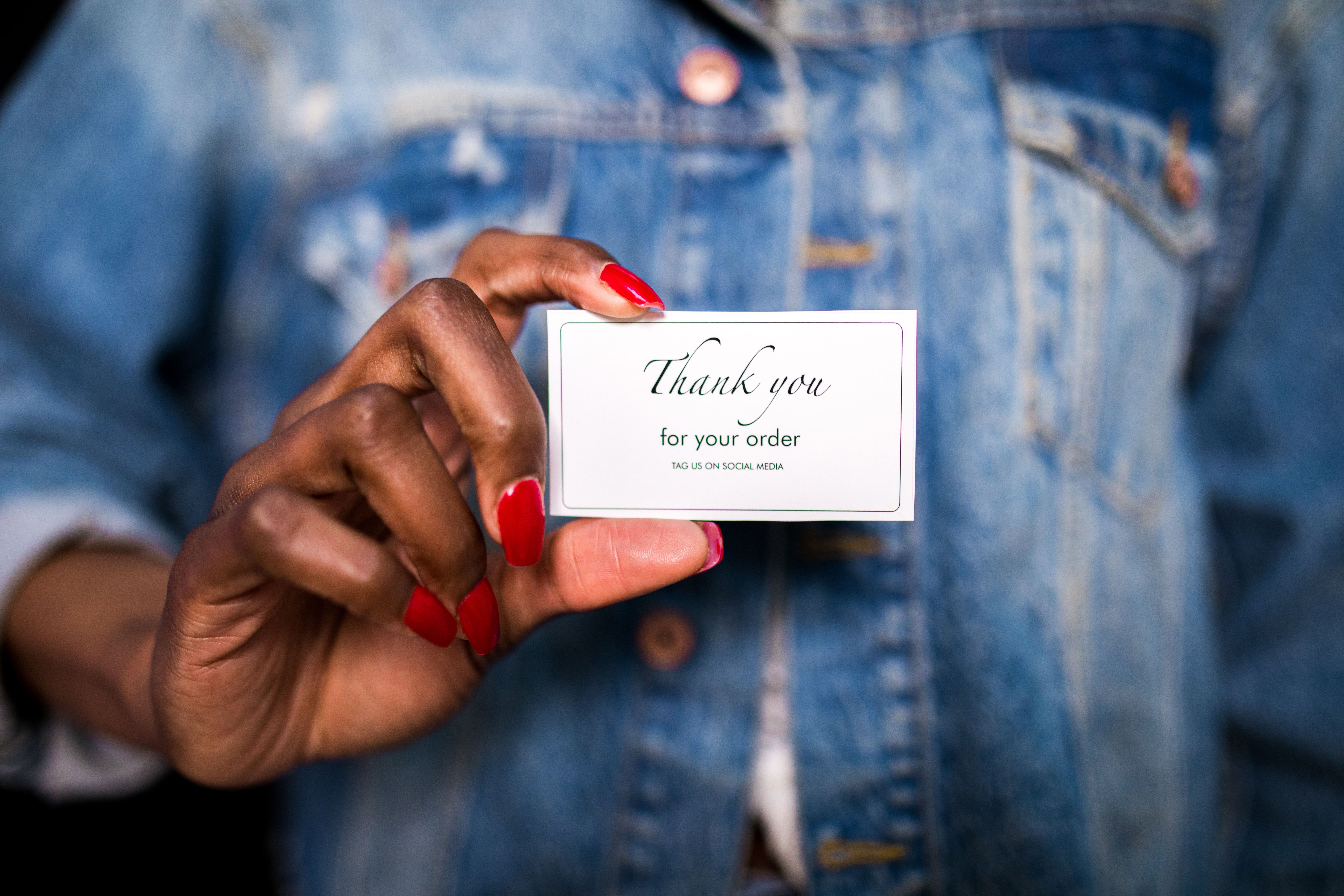 A woman in a denim jacket holds a 'Thank You' card with red nails.